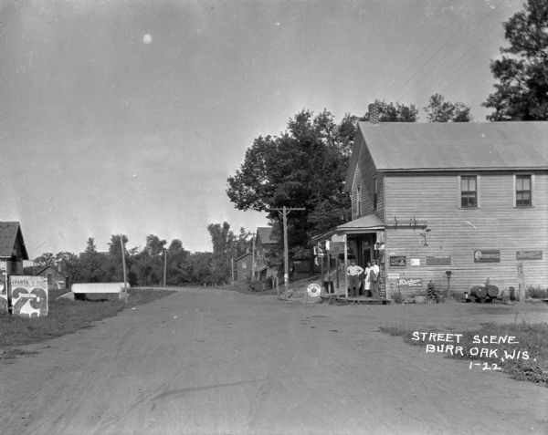 View down road towards three workers standing on the porch of an auto repair shop. The shop offers Red Crown Gasoline, Peerless Motor Oil, and Firestone tires. Across the street, there is an advertisement for the Patterson's Circus show in Sparta on June 29th.