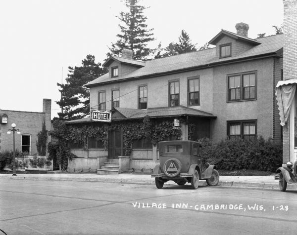 View across road of the Village Inn; featuring a "Hotel" sign, vines along the screened porch windows, and a fenced garden.