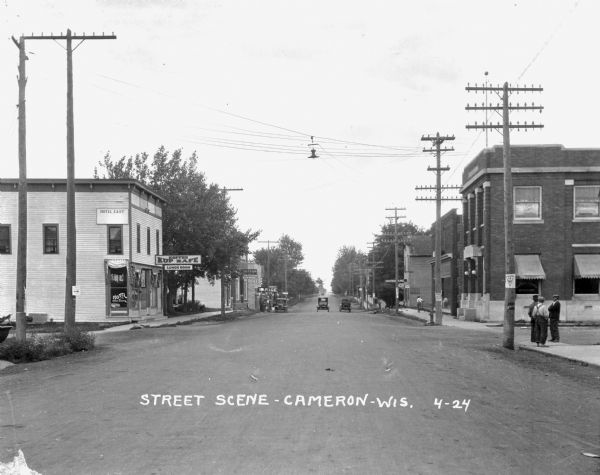 A view down a street with stores and shops. The stores include: Koffee Kup Kafe, Hotel East, Boortz Garage, a hardware store, and the State & County Depository. Three men gather at a street corner on the right, and further down the sidewalk a young boy pushes a reel mower.