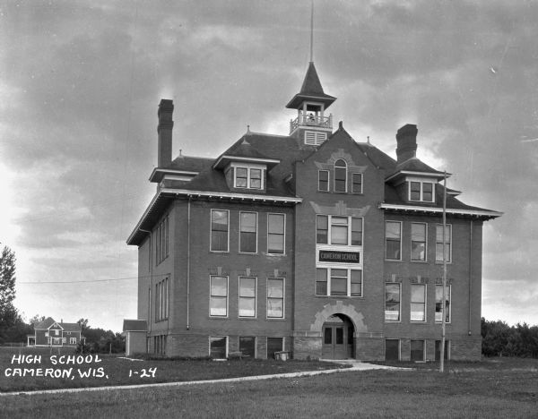 Exterior view of the Cameron High School. The building features three stories, a bell tower, and a flag pole. A home is in the distance.