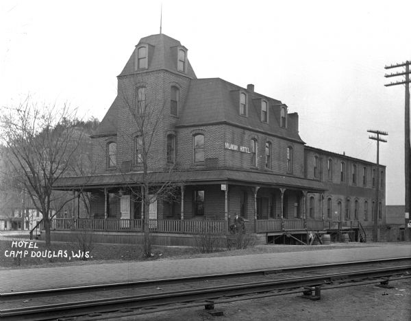 Exterior view over railroad tracks of the Mlakar Hotel near a train station. A man sits on the porch of the hotel.