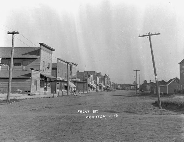 View down Front Street of stores and shops. At the far end of the street is a group of men with horse-drawn wagons.