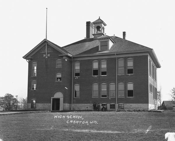 Exterior view of the Cashton High School with bell tower. A woman looks out the window above the entrance, and a group of children and teenagers pose along the wall on the lawn.