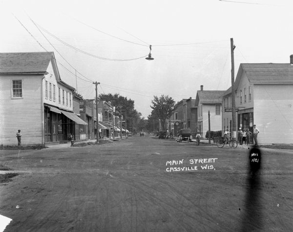 View down middle of commercial street. On the street corner on the right, a man man and children pose on the sidewalk near the drugstore. One of the boys is riding a bicycle. Cars are parked along the curb, and a man is driving a team of horses up the middle of the street in the background.