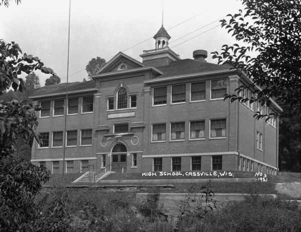 Exterior of the Cassville High School. The building features a fanlight window, bell tower, widow's walk, and two flagpoles; one on top and one in front of the building.