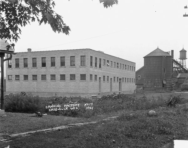 The back of the Klindt-Geiger Canning Company grounds. The site includes a two-story factory, a silo, and a water tower. There is a reel lawn mower lying on the grass.