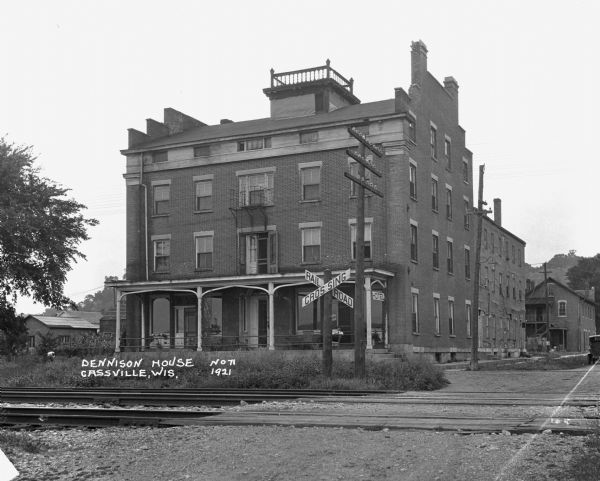 View across railroad crossing of the Dennison House hotel. A man is sitting on the porch. The building features a widow's walk. There is a bluff in the far background.