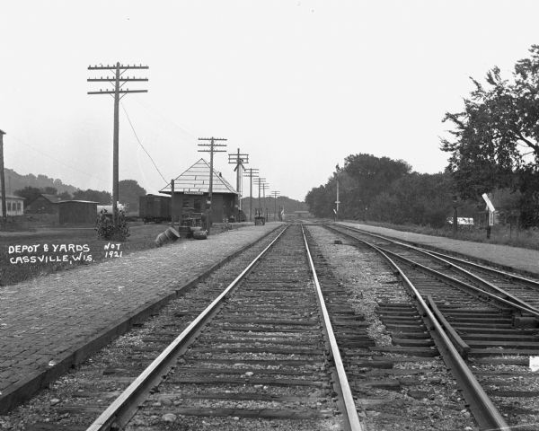 View down railroad tracks of the Cassville depot. A boy or man sits on a bench in front of the depot.