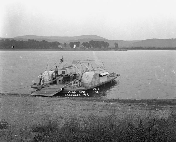View from shoreline looking down at three men posing on a paddle steamer ferry boat carrying an automobile. The boat has the words "Cassville" and "Dewey" painted on it. There are buildings on the far shoreline.