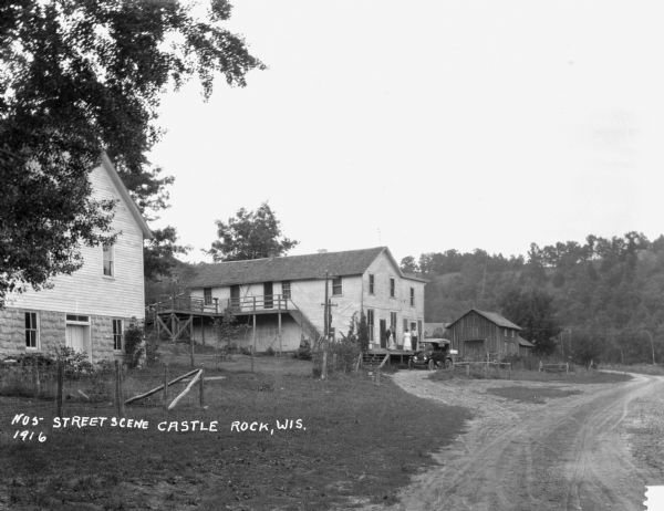 View down driveway of two men and a woman stand on the open porch or loading dock of a building. A car is parked next to the group. There are other buildings nearby. A bluff is in the background.