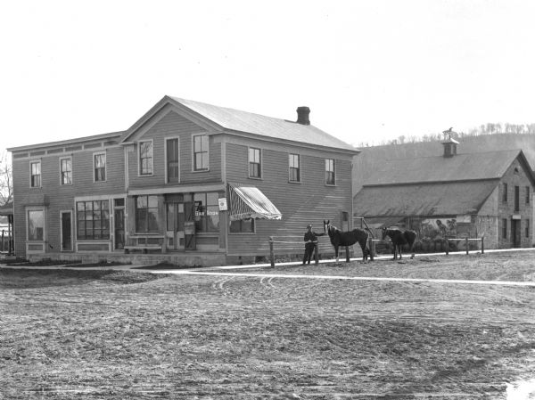 View across road of a man with his horse outside the Bar Room. There is a large circus advertisement with elephants and lions painted on the wall of the building behind the bar. A small sign near the front window says, in part, "Lager Beer" and possibly "Fauerbach." There is a bluff in the background.