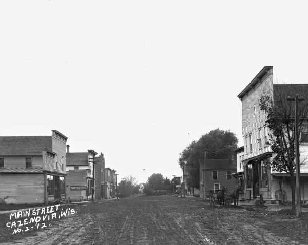 View down Main Street. A team of horses pulling a buggy is parked at the curb near a storefront on the right. There is a shoe store and hotel on the left.