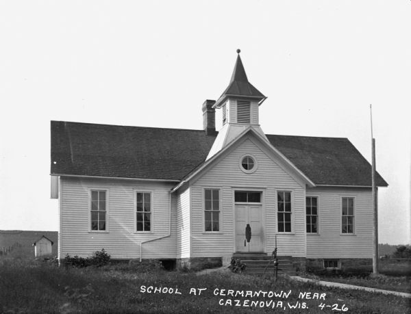 Exterior of a schoolhouse with a hand-pump water well and flagpole in front.