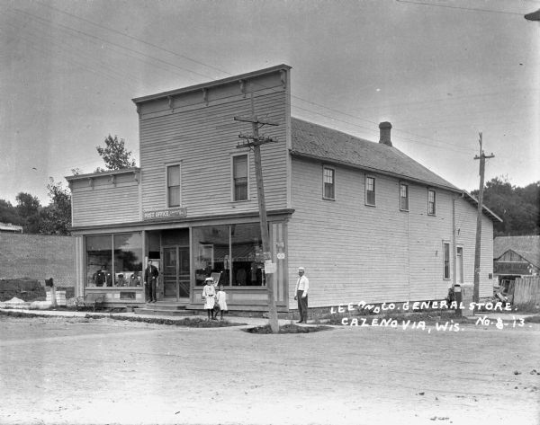 View from road of the exterior of the Lee and Company General Store/post office. A young man stands on the porch; two girls walk on the sidewalk, and a man stands at the corner. There is an advertisement for Spear Head tobacco on the shed behind the store.