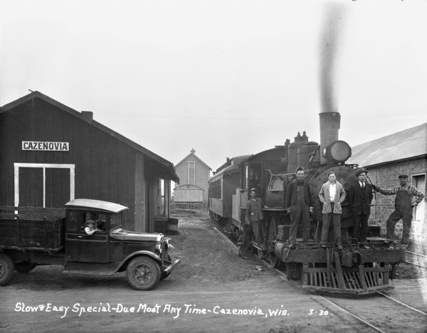 Scene at the Cazenovia train depot. A group of men pose on the front of the locomotive. A man and a child pose from inside a parked pickup truck. Written on negative: "Slow & Easy Special — Due Most Any Time."