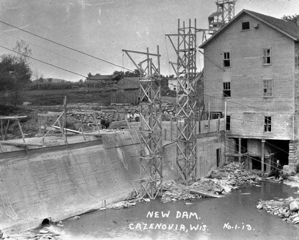 View looking down on construction site of a dam. Two pulley-operated hoists are erect. Men pose from the top of the dam.