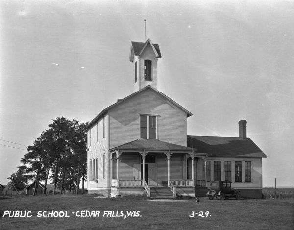 Exterior view of the public school. The school building features two stories, a bell tower, and a porch. A car is parked by the entrance. There is a swing set in the yard to the left of the school.