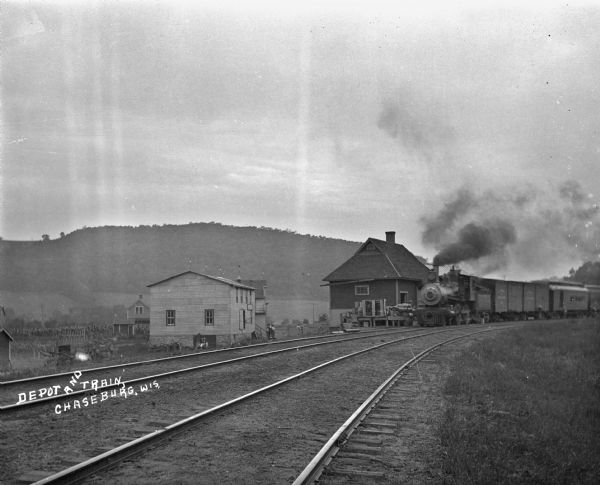Train departing the Chaseburg depot, with steam coming out of the smokestack. There are buildings along the tracks, and in the background is a bluff.