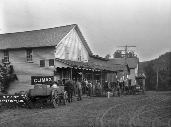 View from road of men and women posing at the entrance of a general store. Horses and wagons are at the curb. The store advertises Jewel paints, Climax tobacco, and Horse shoe tobacco.