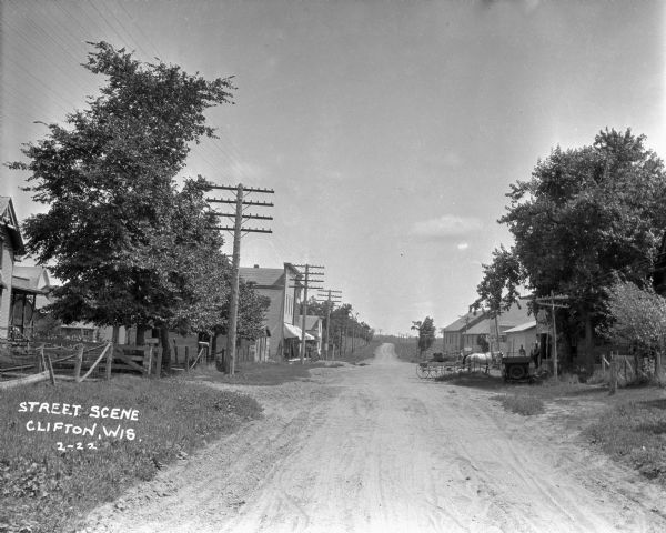 View down an unpaved street in a residential area, and storefronts further down. Near a garage on the right is a horse-drawn wagon and automobile. Two men and a young boy stand nearby in front of a building.