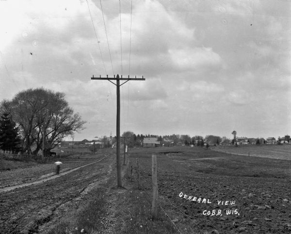 View along fence line of a rural area. There is a road on the left and farmland on the right. A person is traveling down the road, visible as a blur. In the far background is a town.