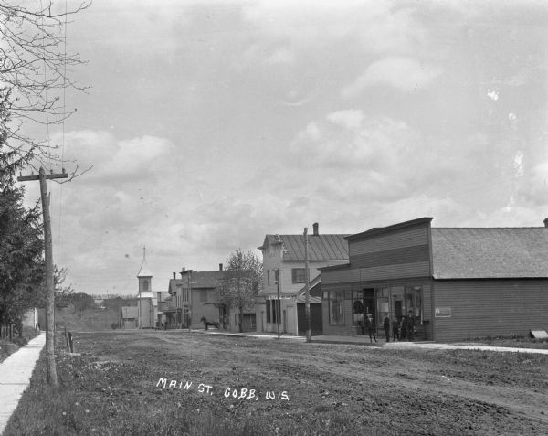 View across Main Street, with a clothing store, livery stable, bank, and church on the right. A group of young men and boys stand on the sidewalk wearing suits and hats.