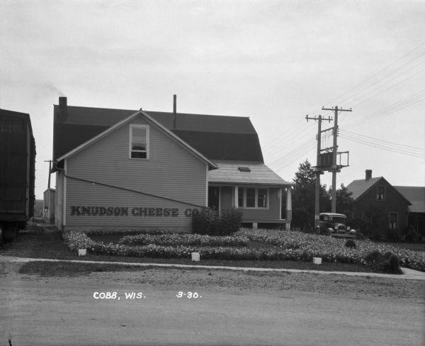 Exterior of the Knudson Cheese Company. The lawn is covered with flowers. A man sits on the porch. A railroad car is on the left.