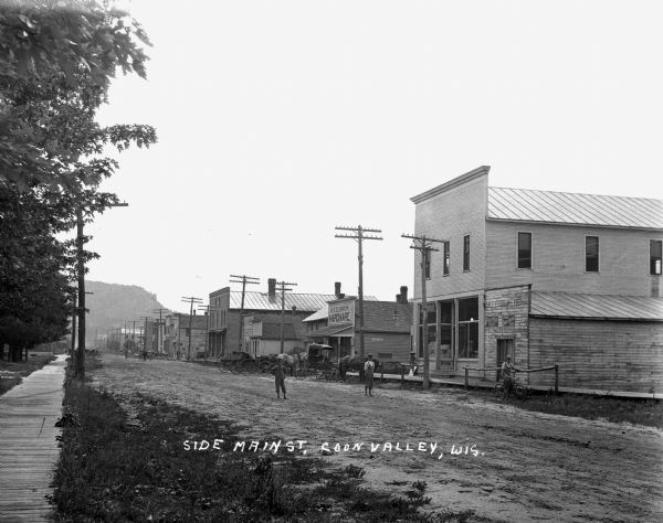 Two boys, barefoot and in overalls, stand in the middle of the unpaved street. A boy on a fence and a man on a bicycle are near the curb. The shops on the street are O.P. Dunnum Hardware and a general store. There are several horse-drawn buggies at the curb.