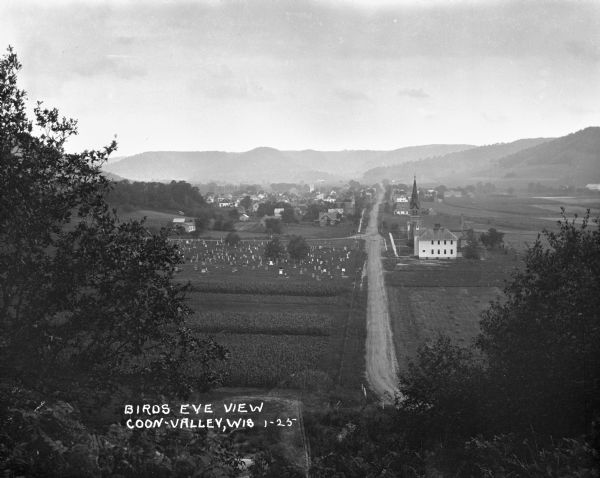 Elevated view of Coon Valley from a hilltop. Trees are on either side of the frame. In the city, there is a cemetery, church, and field of crops.