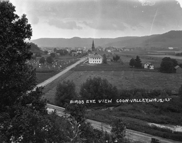 View of a church, field, and cemetery from a hilltop. A car drives down the road in the lower-right corner.