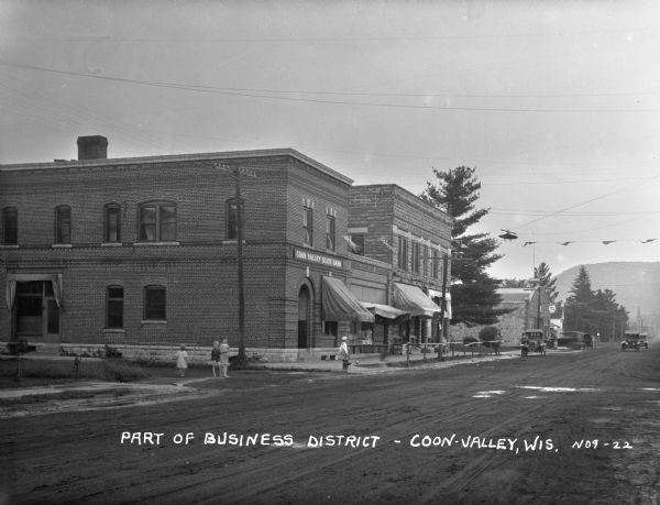 View across street of children on the sidewalk outside the Coon Valley State Bank. At the end of the street, there is an automotive repair station.