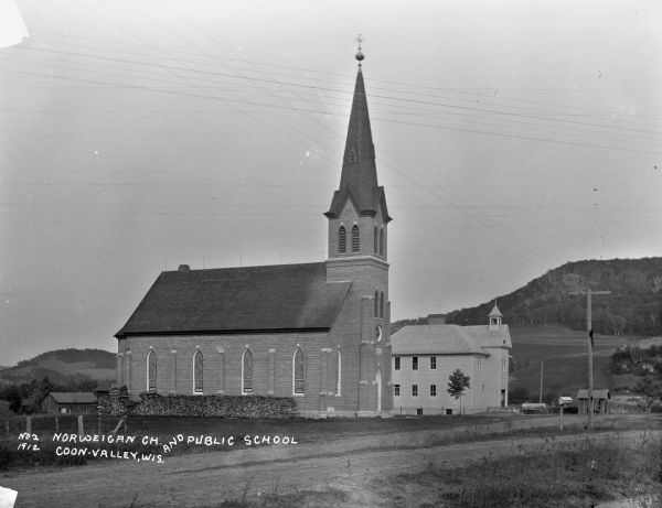View across road of a Norwegian Church, and a Public School next door. The church building features a tall steeple, lancet arched windows, gable roof, and  a rose window. There is fuelwood stacked along the churches' wall.