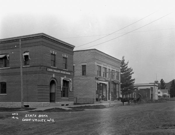 View from across the intersection of Coon Valley State Bank, D.O. Stevlingson - the Gotzian Shoe, and the Restaurant Colonial. A horse and buggy is tied at the curb of the restaurant.