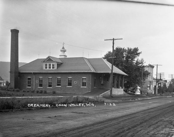 Exterior view across road of a creamery. The front of the creamery has a roof supported with columns over the elevated driveway and loading dock for deliveries. A tall chimney is on the left near the rear of the building, and a large metal roof vent with weathervane is in the middle of the hip roof. In the background is a bluff. A sign for International farm equipment is in front of a building on the right.