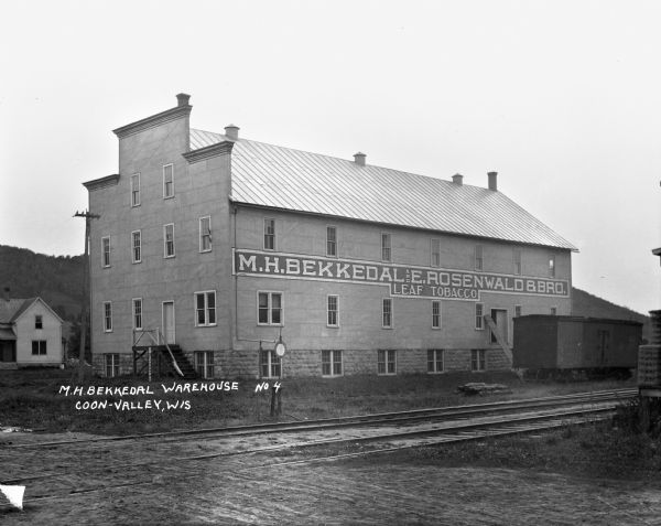 M.H. Bekkedale and E. Rosenwald and Brothers Leaf Tobacco warehouse. There is a boxcar on tracks near the warehouse, and a bluff in the far background.