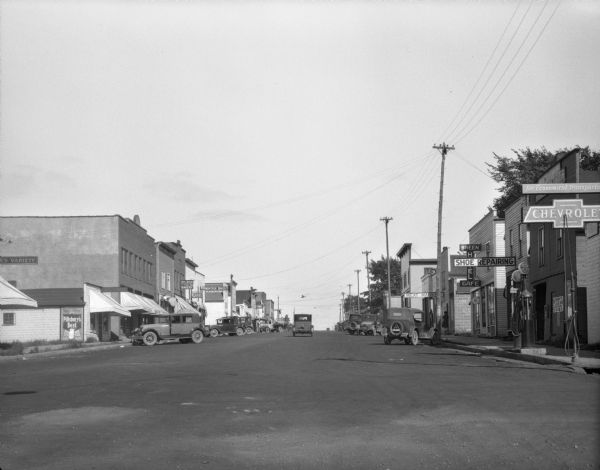 A busy street scene. Cars are parked at the curbs along the shops. The shops include: a grocery, a bakery/ice cream shop, a cafe, Prentice brothers hardware store, a drugstore, a meat market, Breen Hotel and Cafe, Shoe repair shop, and a Chevrolet dealership.