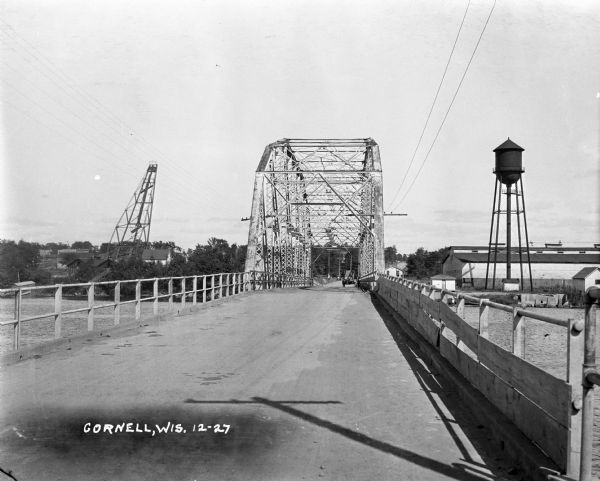 View down middle of metal bridge across the Cornell Flowage. Across the lake, there is the pulpwood stacker on the left and a water tower on the right near factory buildings.