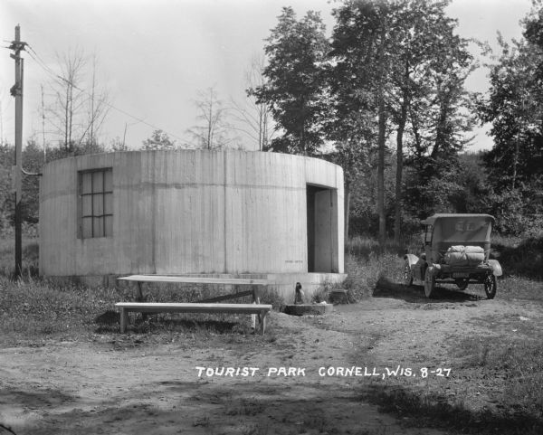 A cylinder-shaped cement building with an open door in a rural area. In front of the building is a picnic table, and on the right an automobile is parked nearby. Near the doorway is a pipe with water running into a concrete basin. The words "spring-water" are on the wall above it.
