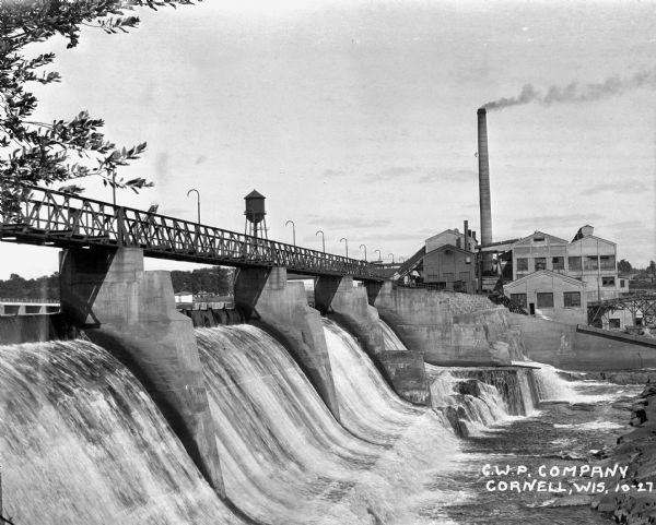 View across lower section of dam at Cornell Wood Products Company. In the background are factory buildings, a tall smokestack, and a water tower.