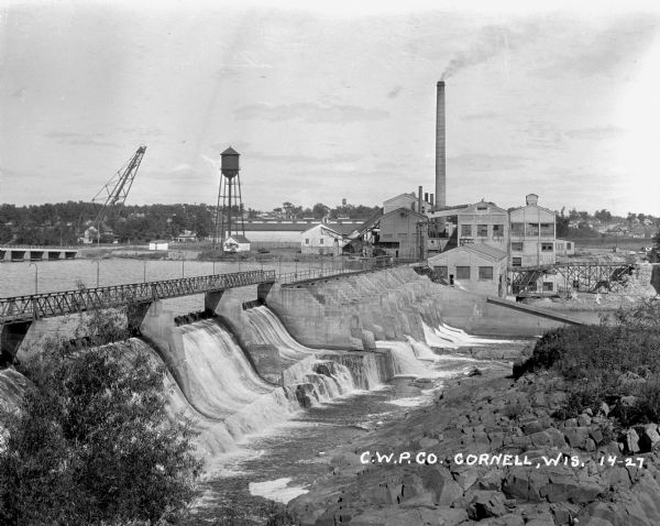 Elevated view over dam of the site of the Cornell Wood Products Company. On the opposite shoreline is a pulpwood stacker, tall smokestack, water tower, and factory buildings.