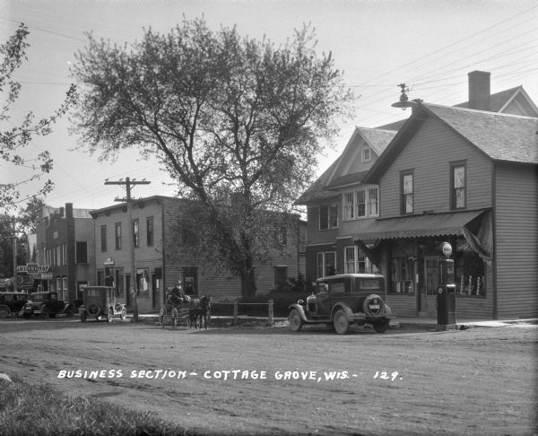 View across commercial street looking south. Parked at the curb are cars, and driving along the street is a bearded man with a horse and buggy. The shops are: Sime B. Barron grocery shop and gas station, a post office (?), and a Chevrolet/Pontiac repair shop and gas station.