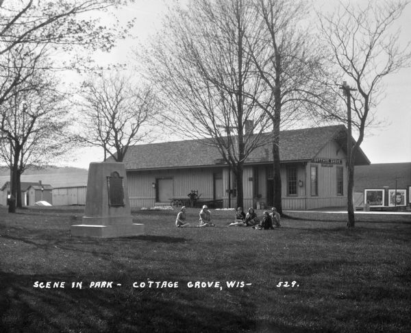 A group of young women sit on the grass at a park. There is a monument in the park. A man stands near the Cottage Grove train depot in the background.