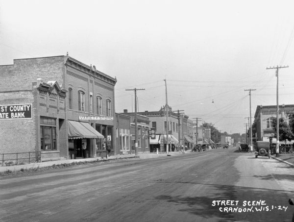 View down commercial street. The stores are (from left to right): First County State Bank, a variety store, a bakery, First National Bank, a drugstore, the Hotel Hellstrom, a restaurant, and another drugstore. Cars are parked at the curbs, and pedestrians are on the sidewalks.