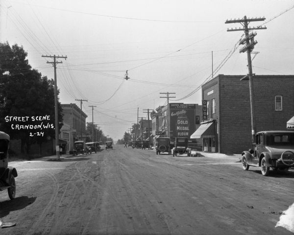 View down commercial street. The shops are: Crandon Baking Company, a drugstore, the Hotel Hellstrom and Livery, a restaurant, First National Bank, a bakery, and a variety store. There is a large Gold Medal Flour advertisement painted on the side of the Hotel.