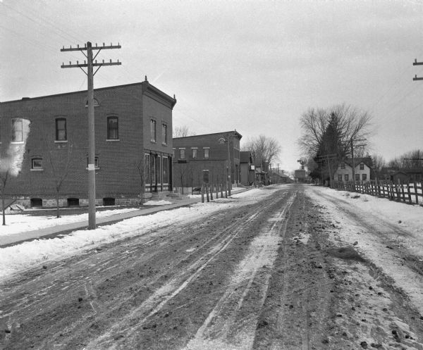 View down a snowy unpaved street. There appears to be a water tower, as well as a windmill at the far end of the street.