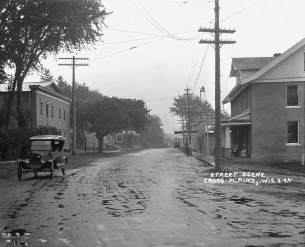 View down a rainy commercial street. There is a soda/ice cream shop, garage/gas station, and undertaker. A car is parked on the side of the road. There is a bell on a tall tower behind buildings on the right.