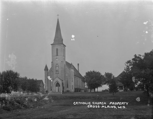 Exterior view of Catholic Church. The building features a tall steeple, a clock tower, arched, stained-grass windows, and double doors.