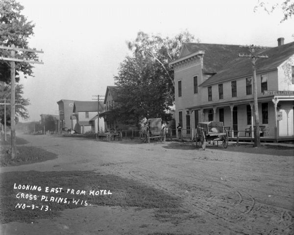 Exterior view of a hotel and livery from across an unpaved road. Three horse-drawn vehicles are tied at the curb.