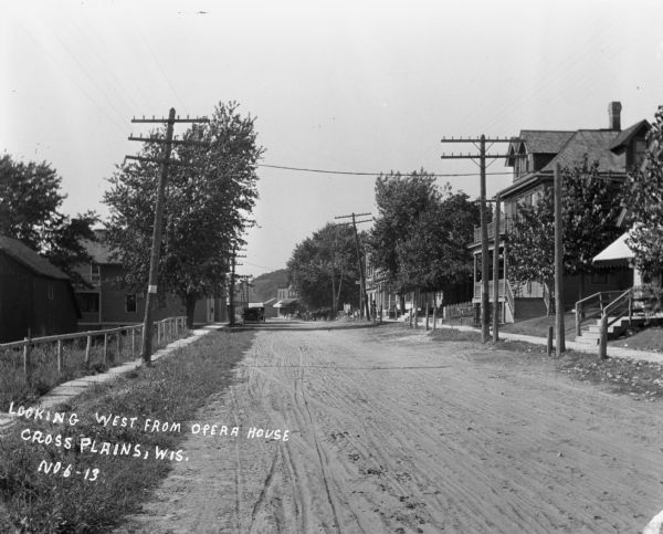 View down an unpaved street. There are homes on the right and a fence on the left. At the end of the street are commercial buildings, horses, and automobiles. A bluff is in the far background.