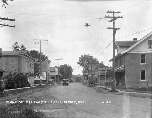 View down a commercial street, with an automobile coming down the street. There is a soda/ice cream shop which offers tobacco, cigars, and candy. The City Garage advertises Goodyear Tires, Mobiloil oil, Chevrolet and Essex parts, and its AAA affiliation. On the other side of the street there is an undertaker/funeral home. A truck is parked on the right near the garage.